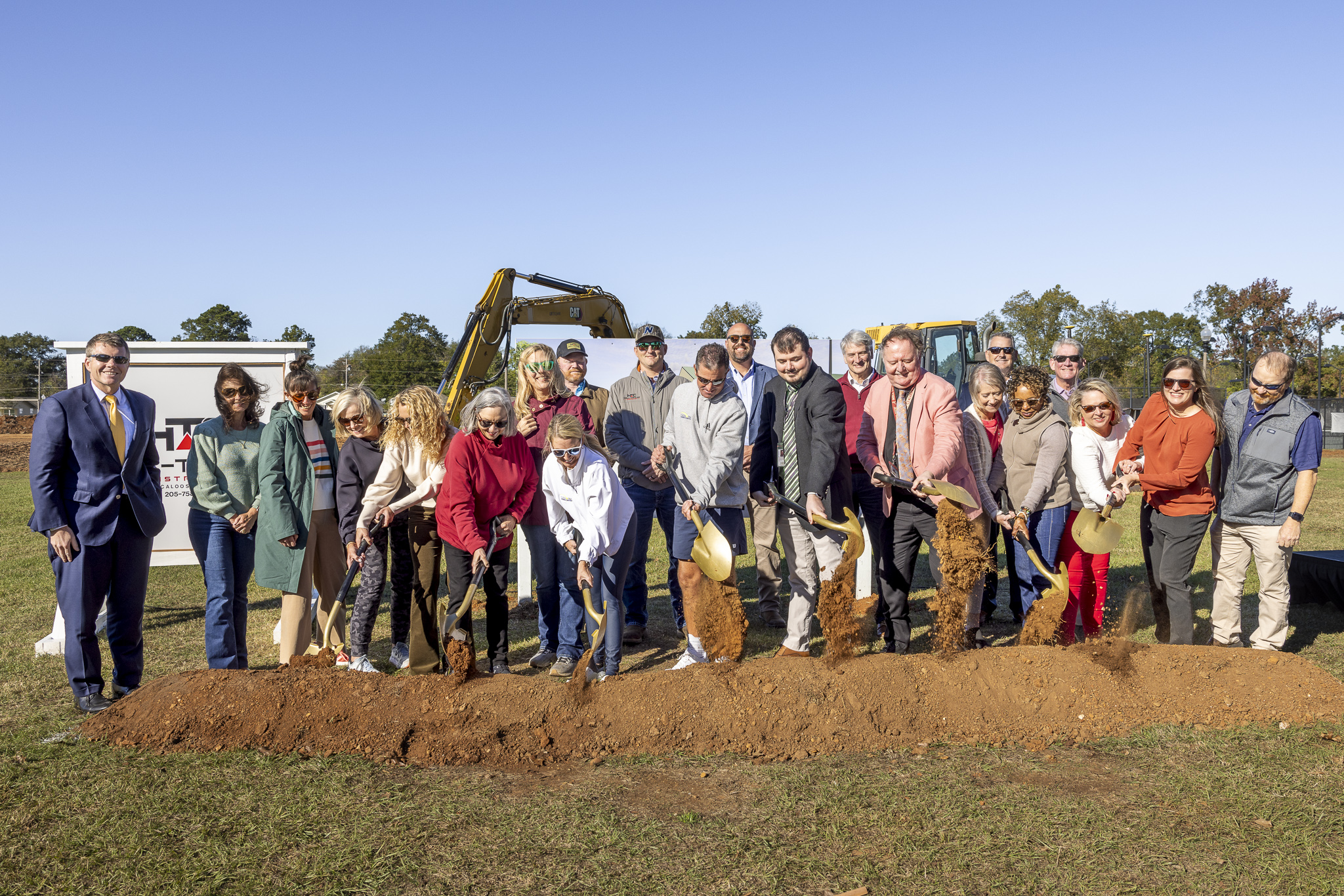 A large group of people smiles behind a dirt pile with shovels in the dirt.